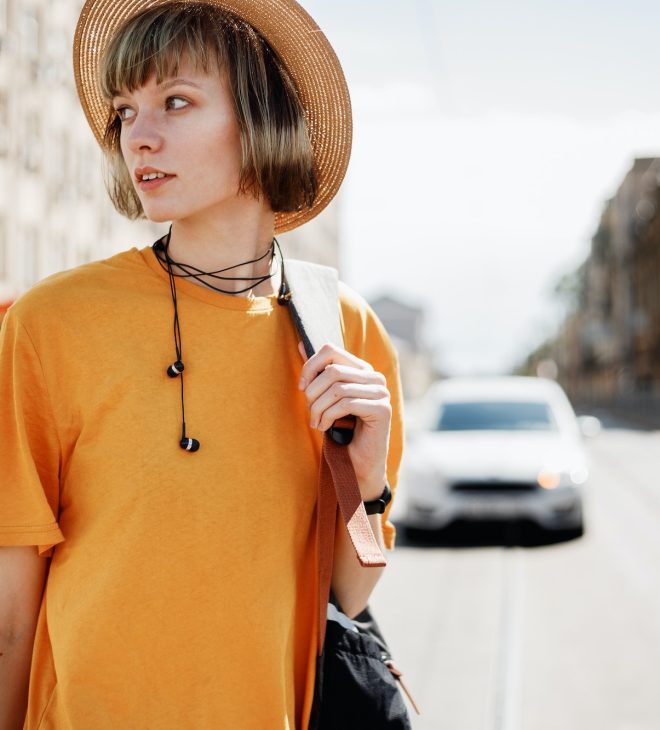 Young girl with headphones in a yellow T-shirt and a straw hat walks with a backpack along a city